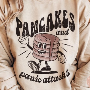 May include: A beige sweatshirt with a graphic design of a smiling pancake character with the text "Pancakes and panic attacks".