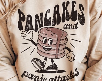 Puede incluir: Una sudadera beige con un diseño gráfico de un personaje de panqueque sonriente con el texto "Pancakes and panic attacks".