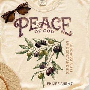 May include: A cream-colored t-shirt featuring a graphic of an olive branch with dark and green olives. The text reads PEACE OF GOD, SURPASSES ALL UNDERSTANDING, and PHILIPPIANS 4:7. Tortoiseshell sunglasses and a straw hat are placed on the shirt.