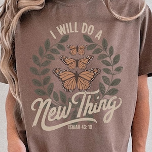 May include: Brown t-shirt with the text "I WILL DO A New Thing" and the Bible verse Isaiah 43:19. The design includes three orange butterflies and green leaves. The shirt is a relaxed fit.