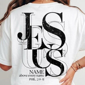 Puede incluir: Una camiseta blanca de manga corta con un gráfico negro desgastado en la espalda que dice JESUS en letras negritas. Debajo del texto principal, una letra negra más pequeña dice NAME above every name y la referencia bíblica PHIL. 2:9-11.