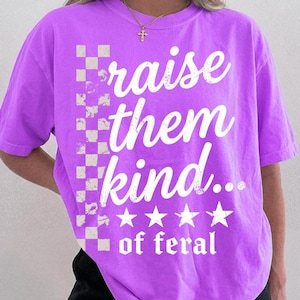 Pode incluir: Uma t-shirt de manga curta roxo brilhante com uma impressão gráfica branca que diz "raise them kind... of feral", um padrão xadrez vertical no lado esquerdo e quatro estrelas brancas acima do texto "of feral".