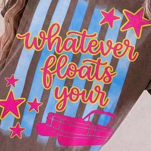 May include: Brown t-shirt with the phrase "whatever floats your" in pink script with a yellow outline. The design includes blue stripes, pink stars, and a pink pontoon boat graphic.