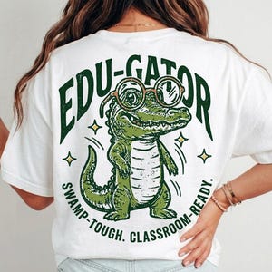 May include: A white t-shirt featuring a green graphic of an alligator wearing round glasses. The text reads EDU-GATOR at the top and SWAMP-TOUGH. CLASSROOM-READY. at the bottom. The design includes small star accents surrounding the alligator.