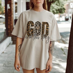 Puede incluir: Una camiseta beige con un dise&ntilde;o de estampado de leopardo que deletrea "God is in this story" con un peque&ntilde;o coraz&oacute;n en el medio de la palabra "God".