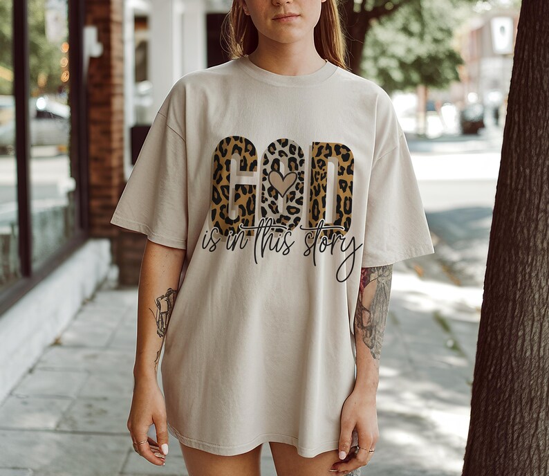 Puede incluir: Una camiseta beige con un dise&ntilde;o de estampado de leopardo que deletrea "God is in this story" con un peque&ntilde;o coraz&oacute;n en el medio de la palabra "God".