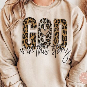 Puede incluir: Una sudadera beige con un dise&ntilde;o de estampado de leopardo que dice "Dios est&aacute; en esta historia" con un peque&ntilde;o coraz&oacute;n en el medio de la palabra "Dios".