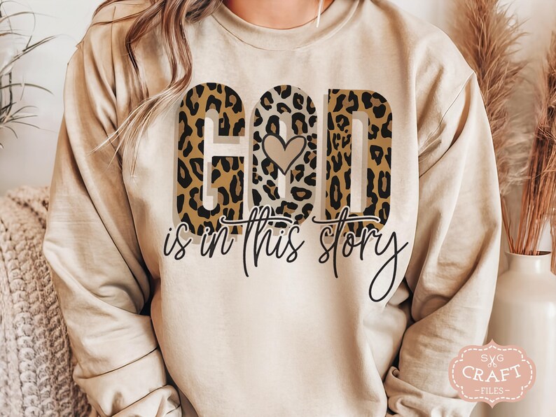 Puede incluir: Una sudadera beige con un dise&ntilde;o de estampado de leopardo que dice "Dios est&aacute; en esta historia" con un peque&ntilde;o coraz&oacute;n en el medio de la palabra "Dios".
