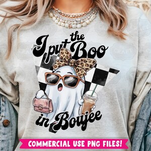 May include: A gray t-shirt with the text "I put the Boo in Boujee" in black and white lettering. A cartoon ghost wearing sunglasses and a leopard print headband is holding a pink purse and a cup of coffee. The background is a black and white checkered pattern.