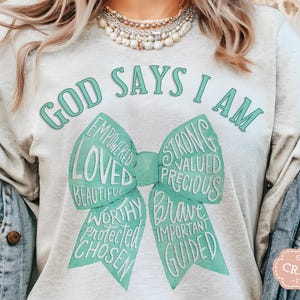 May include: A light gray t-shirt with the text "GOD SAYS I AM" in teal. A teal bow graphic features words like "Loved", "Strong", and "Brave". The shirt is a casual style.