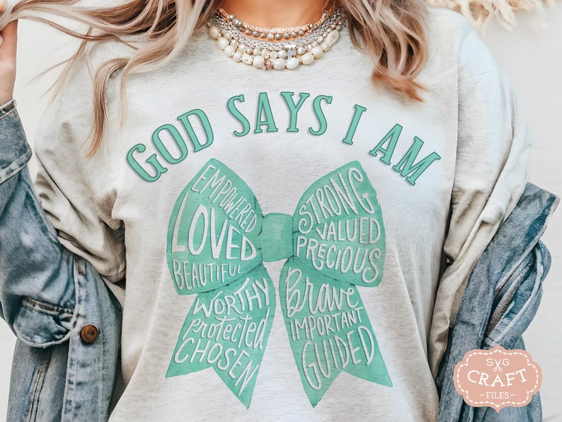 May include: A light gray t-shirt with the text "GOD SAYS I AM" in teal. A teal bow graphic features words like "Loved", "Strong", and "Brave". The shirt is a casual style.