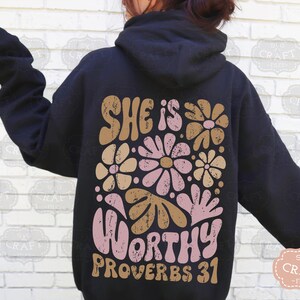 Puede incluir: Una sudadera con capucha negra con un dise&ntilde;o floral y el texto "She is Worthy Proverbs 31" en rosa y marr&oacute;n.