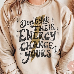 May include: A beige sweatshirt with a black graphic print that reads "Don't Let Their Energy Change Yours" with a gold lightning bolt design.