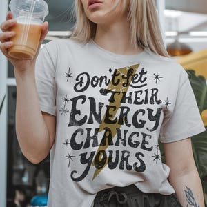 May include: White t-shirt with a black graphic that says "Don't Let Their Energy Change Yours" with a gold lightning bolt design.