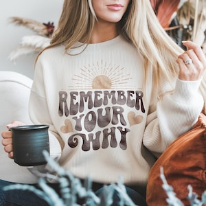 May include: A white waffle knit sweatshirt with a brown graphic that says "Remember Your Why" with a sun and hearts. The sweatshirt is being worn by a person holding a black mug.