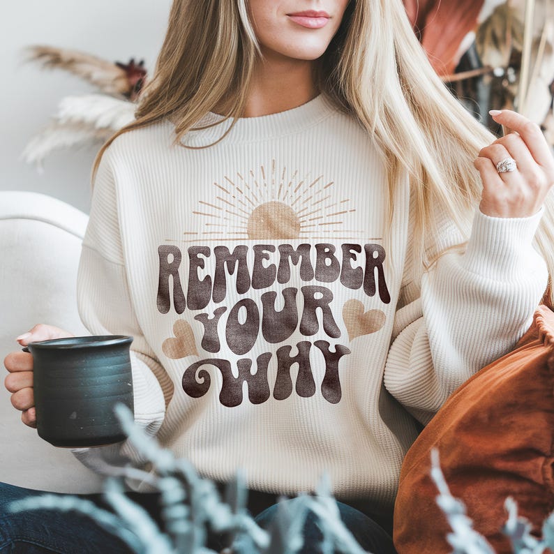 May include: A white waffle knit sweatshirt with a brown graphic that says "Remember Your Why" with a sun and hearts. The sweatshirt is being worn by a person holding a black mug.