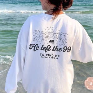 May include: White long-sleeve sweatshirt with a black graphic design of a landscape with a sheep and the text "He left the 99 to find me Matthew 18:12-14".