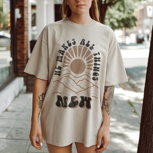 Pode incluir: Uma camiseta bege com uma estampa gr&aacute;fica de um sol e montanhas. O texto "He makes all things new" est&aacute; impresso em preto na camiseta.