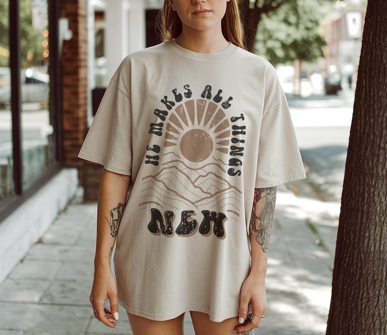 Pode incluir: Uma camiseta bege com uma estampa gr&aacute;fica de um sol e montanhas. O texto "He makes all things new" est&aacute; impresso em preto na camiseta.