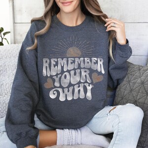 May include: A dark gray sweatshirt with a vintage-style graphic that says "Remember Your Why" in white with a sun and hearts.