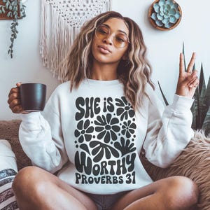 Puede incluir: Sudadera blanca con un dise&ntilde;o gr&aacute;fico negro que presenta el texto "She is Worthy Proverbs 31" rodeado de flores estilizadas.