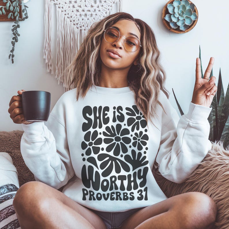 Puede incluir: Sudadera blanca con un dise&ntilde;o gr&aacute;fico negro que presenta el texto "She is Worthy Proverbs 31" rodeado de flores estilizadas.
