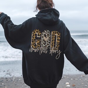 Puede incluir: Sudadera con capucha negra con un dise&ntilde;o de estampado de leopardo que dice "Dios est&aacute; en esta historia" con un coraz&oacute;n en el medio de la palabra "Dios".