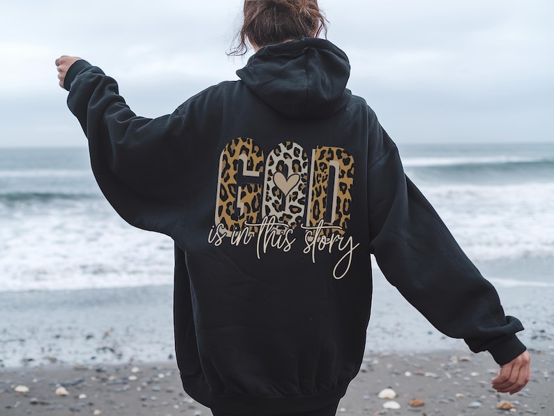 Puede incluir: Sudadera con capucha negra con un dise&ntilde;o de estampado de leopardo que dice "Dios est&aacute; en esta historia" con un coraz&oacute;n en el medio de la palabra "Dios".