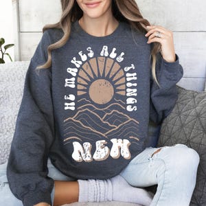 Pode incluir: Uma camisa de moletom cinza escura com um design gr&aacute;fico branco que apresenta um sol, montanhas e o texto "He makes all things new".