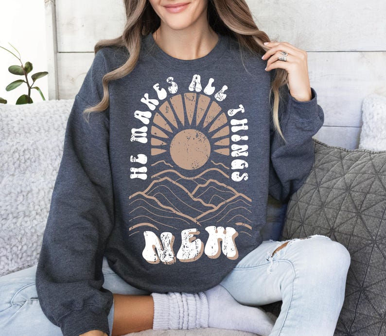 Pode incluir: Uma camisa de moletom cinza escura com um design gr&aacute;fico branco que apresenta um sol, montanhas e o texto "He makes all things new".