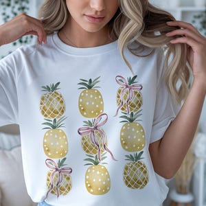 May include: White t-shirt featuring a watercolor design of nine yellow pineapples. Some pineapples have white polka dots, and some are wrapped with pink ribbons. The pineapples have green leafy tops.