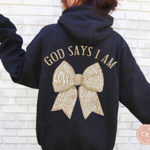 May include: Black hoodie with the text "GOD SAYS I AM" in a gold color. A bow graphic in a tan color is in the center, with words like "Loved", "Strong", and "Brave" inside the bow.
