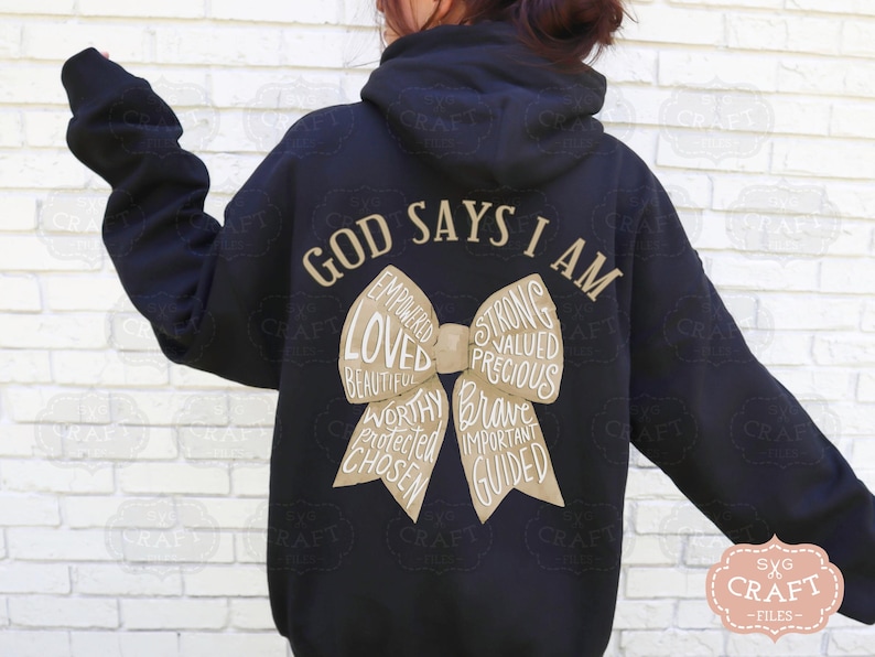 May include: Black hoodie with the text "GOD SAYS I AM" in a gold color. A bow graphic in a tan color is in the center, with words like "Loved", "Strong", and "Brave" inside the bow.