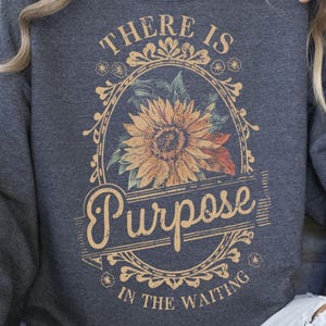 May include: Dark grey sweatshirt with a floral design. The design features a sunflower in an oval frame with the words "THERE IS PURPOSE IN THE WAITING" in a vintage style.