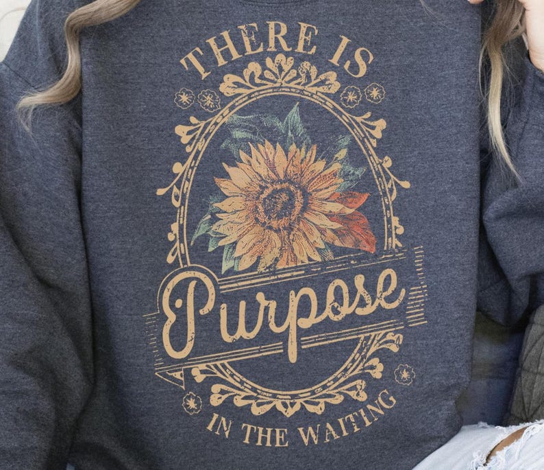 May include: Dark grey sweatshirt with a floral design. The design features a sunflower in an oval frame with the words "THERE IS PURPOSE IN THE WAITING" in a vintage style.