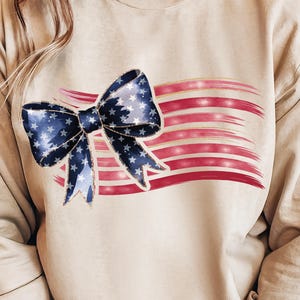 Könnte beinhalten: Ein beiges Sweatshirt mit patriotischem Design. Eine blaue Schleife mit weißen Sternen ist über einem rot-weiß gestreiften Hintergrund zentriert, der einer stilisierten amerikanischen Flagge ähnelt. Die Schleife hat einen goldenen Rand.