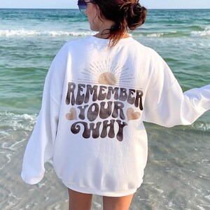 May include: White sweatshirt with a brown graphic that says "Remember Your Why" with a sun and hearts. The sweatshirt is being worn by a person standing on a beach.
