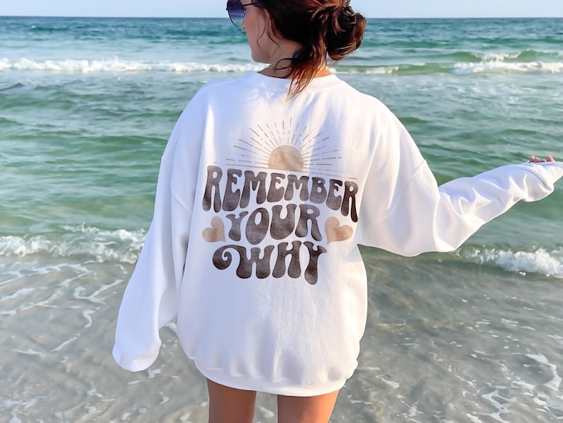 May include: White sweatshirt with a brown graphic that says "Remember Your Why" with a sun and hearts. The sweatshirt is being worn by a person standing on a beach.