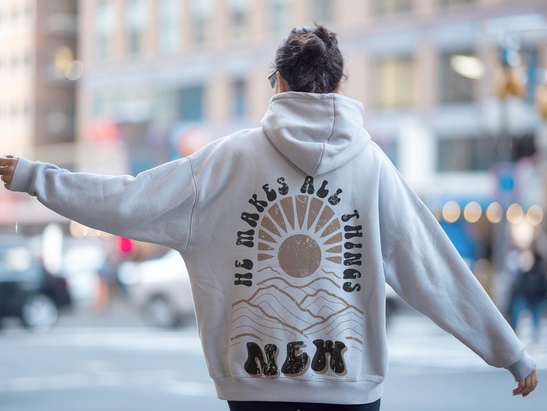 Pode incluir: Uma camisola de capuz cinzenta clara com uma estampa gr&aacute;fica nas costas. O gr&aacute;fico apresenta um sol com raios, montanhas e o texto "He makes all things new".