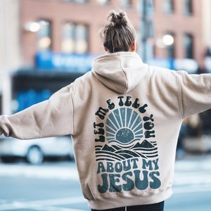 May include: A beige hoodie with a graphic print on the back that says "Let me tell you about my Jesus". The graphic features a blue and white sun with waves and mountains below it.