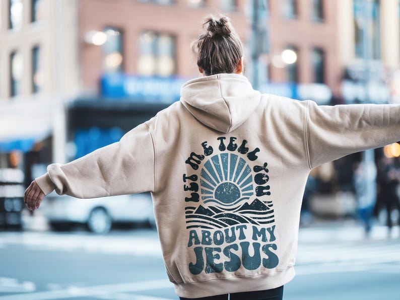 May include: A beige hoodie with a graphic print on the back that says "Let me tell you about my Jesus". The graphic features a blue and white sun with waves and mountains below it.