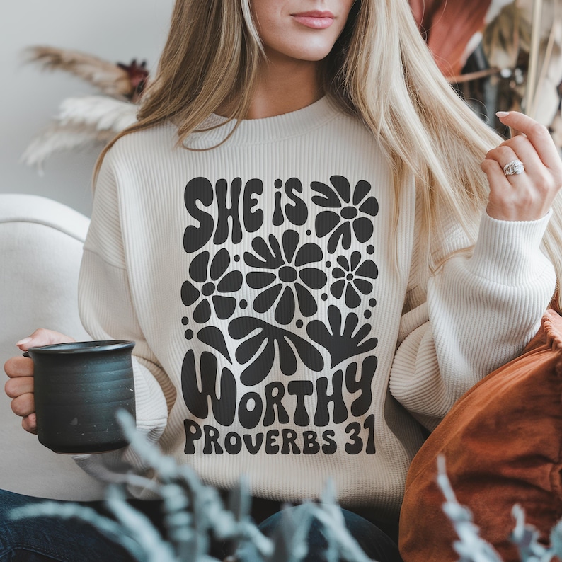Puede incluir: Una sudadera blanca con un dise&ntilde;o gr&aacute;fico negro que dice "She is Worthy Proverbs 31" con un dise&ntilde;o floral alrededor del texto.