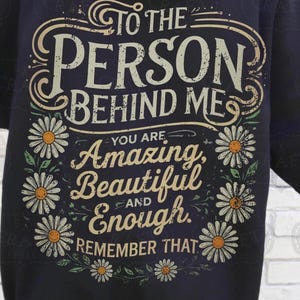 K&ouml;nnte beinhalten: Ein schwarzes T-Shirt mit einem wei&szlig;en Grafikdesign, auf dem "To the person behind me, you are amazing, beautiful and enough. Remember that." steht. Die Grafik ist von wei&szlig;en G&auml;nsebl&uuml;mchen umgeben.