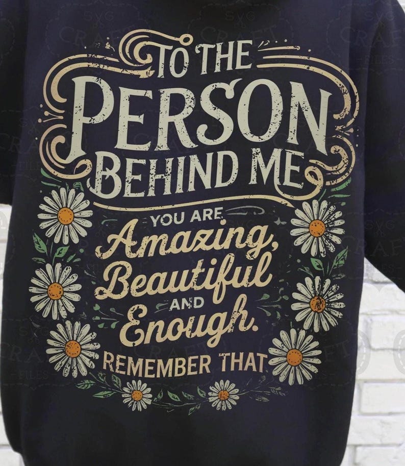 K&ouml;nnte beinhalten: Ein schwarzes T-Shirt mit einem wei&szlig;en Grafikdesign, auf dem "To the person behind me, you are amazing, beautiful and enough. Remember that." steht. Die Grafik ist von wei&szlig;en G&auml;nsebl&uuml;mchen umgeben.