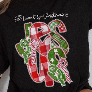 Könnte beinhalten: Schwarzes Sweatshirt mit dem Text "All I want for Christmas is" über einem festlichen "US"-Design. Die Buchstaben sind rot und grün mit Karomustern und Sternen sowie Schleifen.