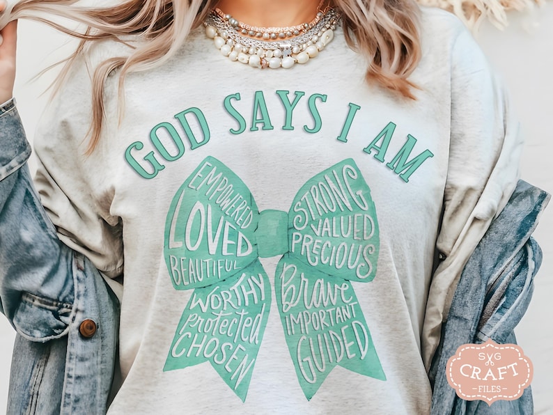 May include: A light gray t-shirt with the text "GOD SAYS I AM" in a teal font. Below the text is a teal bow with words like "LOVED", "STRONG", and "BRAVE". The shirt has a relaxed fit.