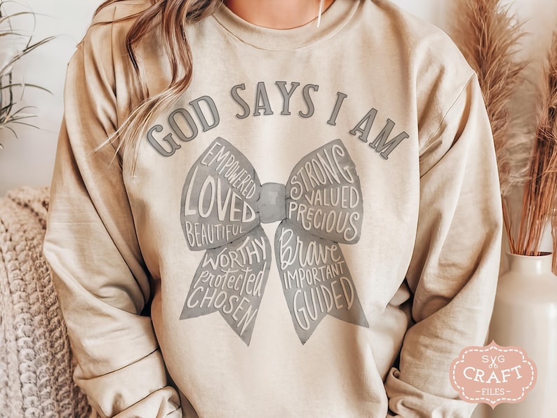 May include: A beige sweatshirt with the text "GOD SAYS I AM" above a bow-shaped graphic. The bow contains words like "LOVED", "STRONG", and "BRAVE". The sweatshirt has long sleeves and a relaxed fit.