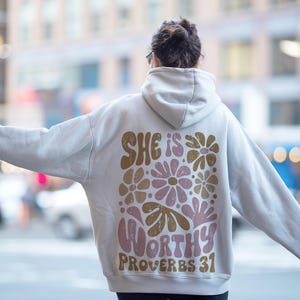 Puede incluir: Una sudadera con capucha blanca con un dise&ntilde;o floral y el texto "She is worthy Proverbs 31" impreso en la espalda.