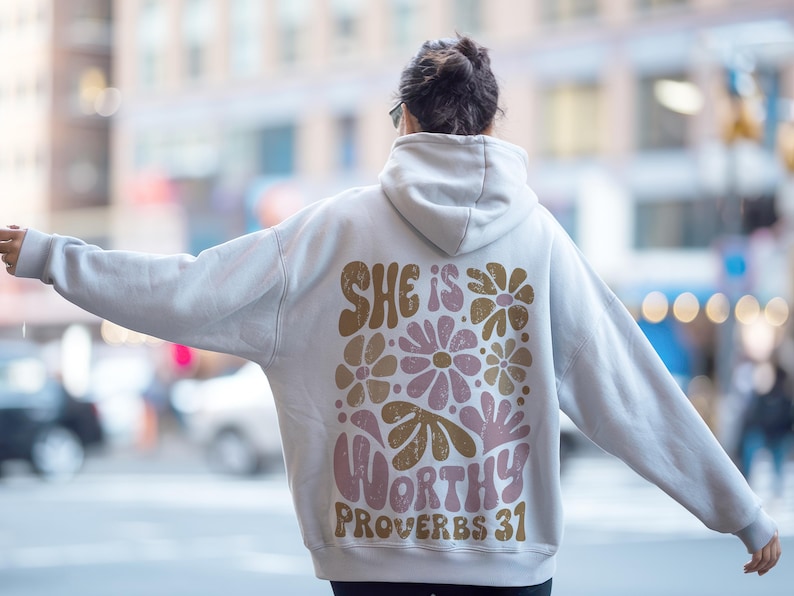 Puede incluir: Una sudadera con capucha blanca con un dise&ntilde;o floral y el texto "She is worthy Proverbs 31" impreso en la espalda.
