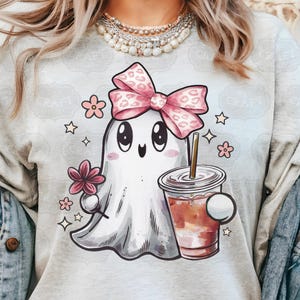 May include: A light gray t-shirt with a cartoon ghost wearing a pink bow and holding a cup of coffee. The ghost has big eyes and a happy expression. The shirt is decorated with small pink flowers and stars.